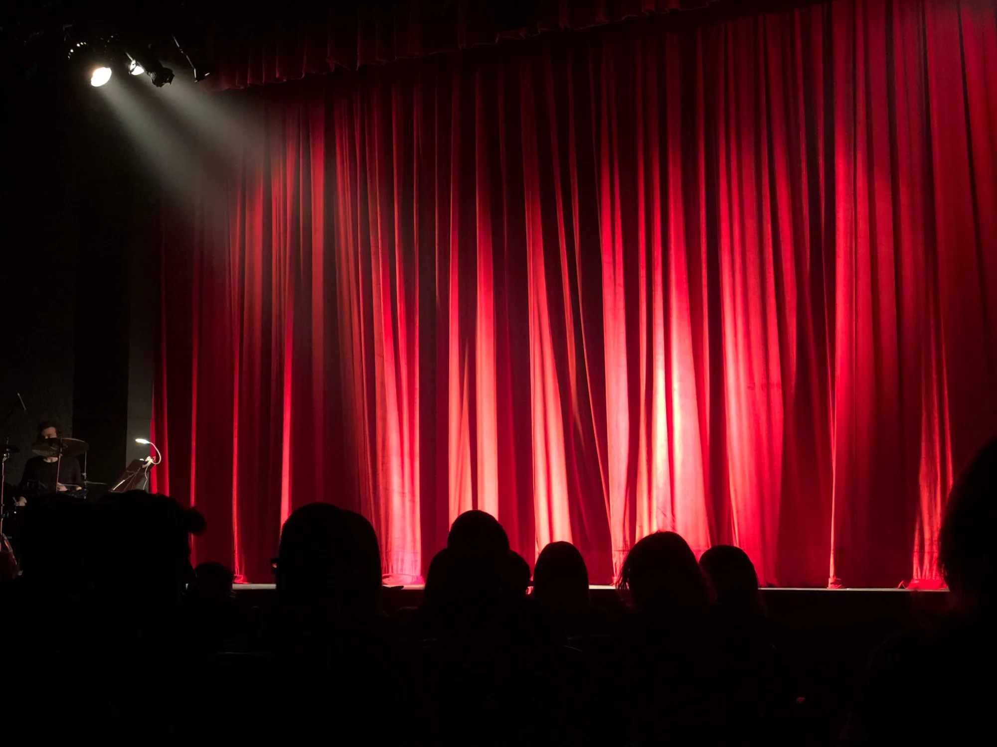 A red theater curtain under a single spotlight, seen from the audience.